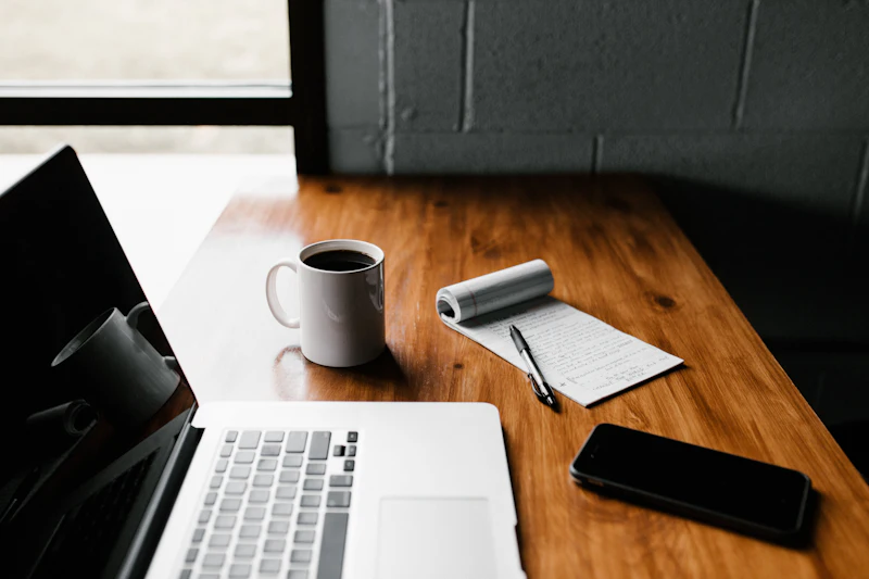 A laptop, a mug and an open notebook on a desk by a window